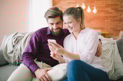 Ein Mann und eine Frau auf einer Couch sehen sich lächelnd etwas auf einem Smartphone an.