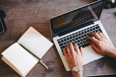 Hände, die auf Laptop-Tastatur tippen, daneben ein Notizbuch