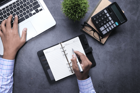 Ein Schreibtisch mit einem Laptop, einem Taschenrechner und einem Notizbuch, in das etwas geschrieben wird