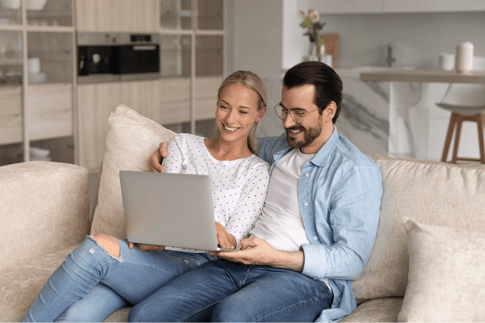 Ein Mann und eine Frau sitzen gemeinsam auf einem Sofa und blicken lächelnd auf einen Laptop
